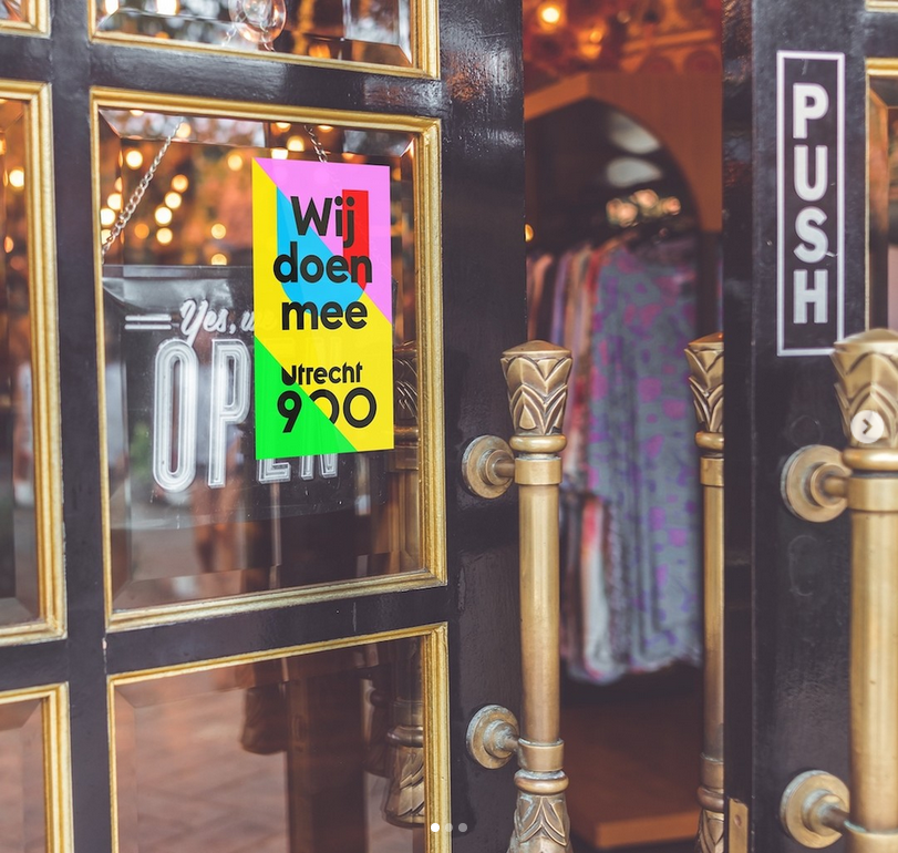 Een poster met de zwarte tekst "Wij doen mee Utrecht 900". De poster hangt op een winkelruit.