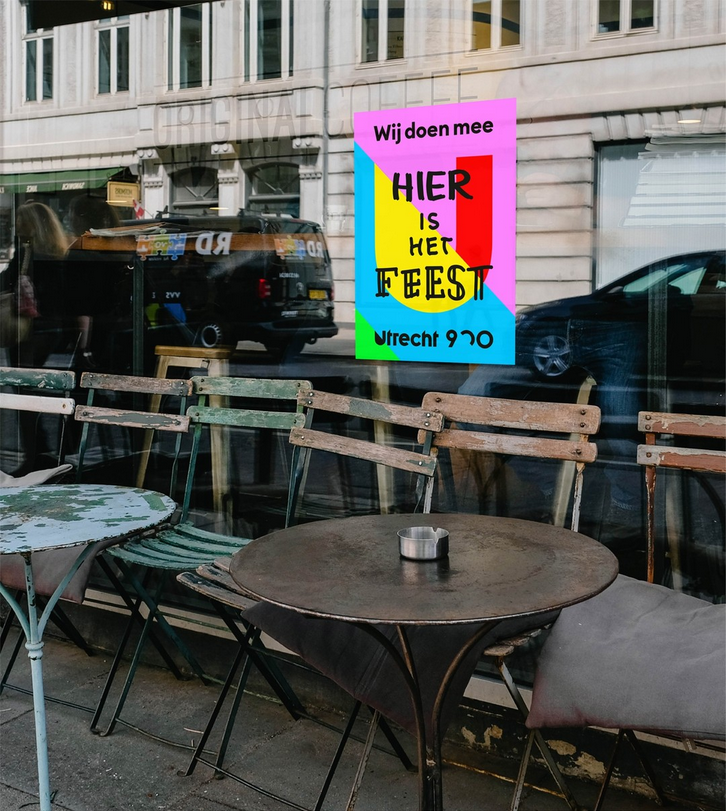 Poster van Utrecht 900 jaar met de zwarte tekst in speelse letters "Hier is het feest Utrecht 900 jaar". De poster hangt in een cafe en op de voorgrond zie je tafeltjes en stoeltjes.